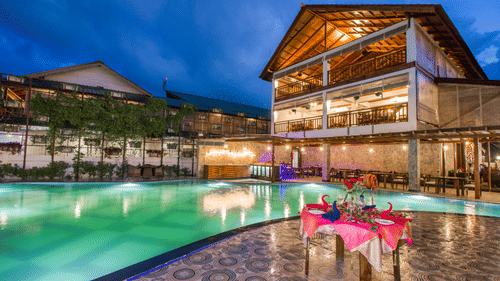 A table next to a swimming pool decorated with pink sheets with the facade of Symphony Summer Sand Beach Resort And Spa, Neil Island at the background.