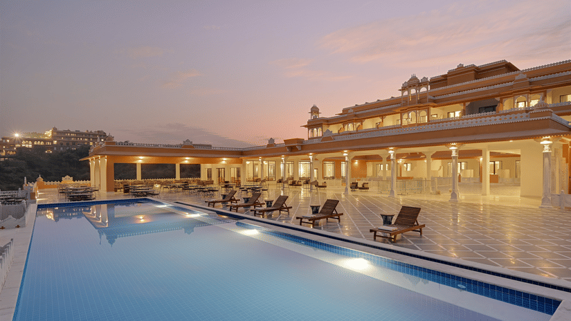 Facade of Fateh Vilas in Udaipur featuring a well lit building and a swimming pool.