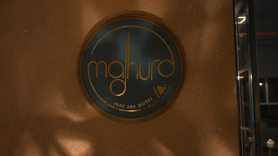 A close up of the Madhura logo displayed on a polished brown surface at Hotel Sonar Bangla, Mayapur.
