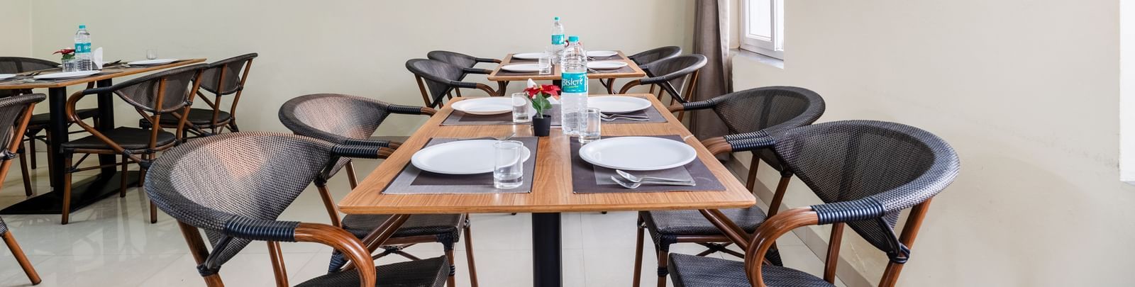 An empty dining area at Destino by Monday Hotels, Gachibowli, with 2 dining tables and woven chairs, set up with plates and water bottles.