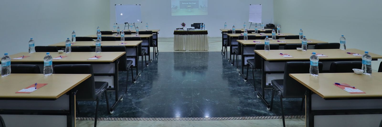 A spacious room with rows of tables and chairs facing a front presentation area with a screen - Retreat Heritage, Lonavala