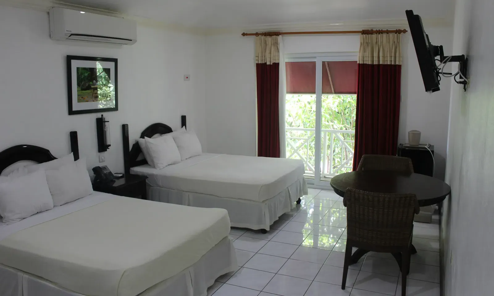 A spacious hotel room contains two beds with white bedding, a dark dresser with a television, and red curtains | Deluxe Double Room | Altamont Court, Jamaica