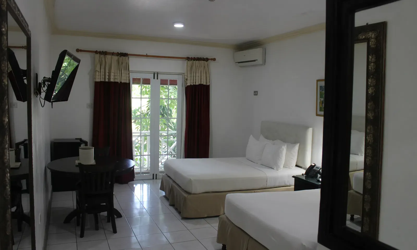 A bright hotel room displays a large bed with white linens, a dark dresser, and a seating area with a table and chairs | Deluxe Double Room | Altamont Court, Jamaica