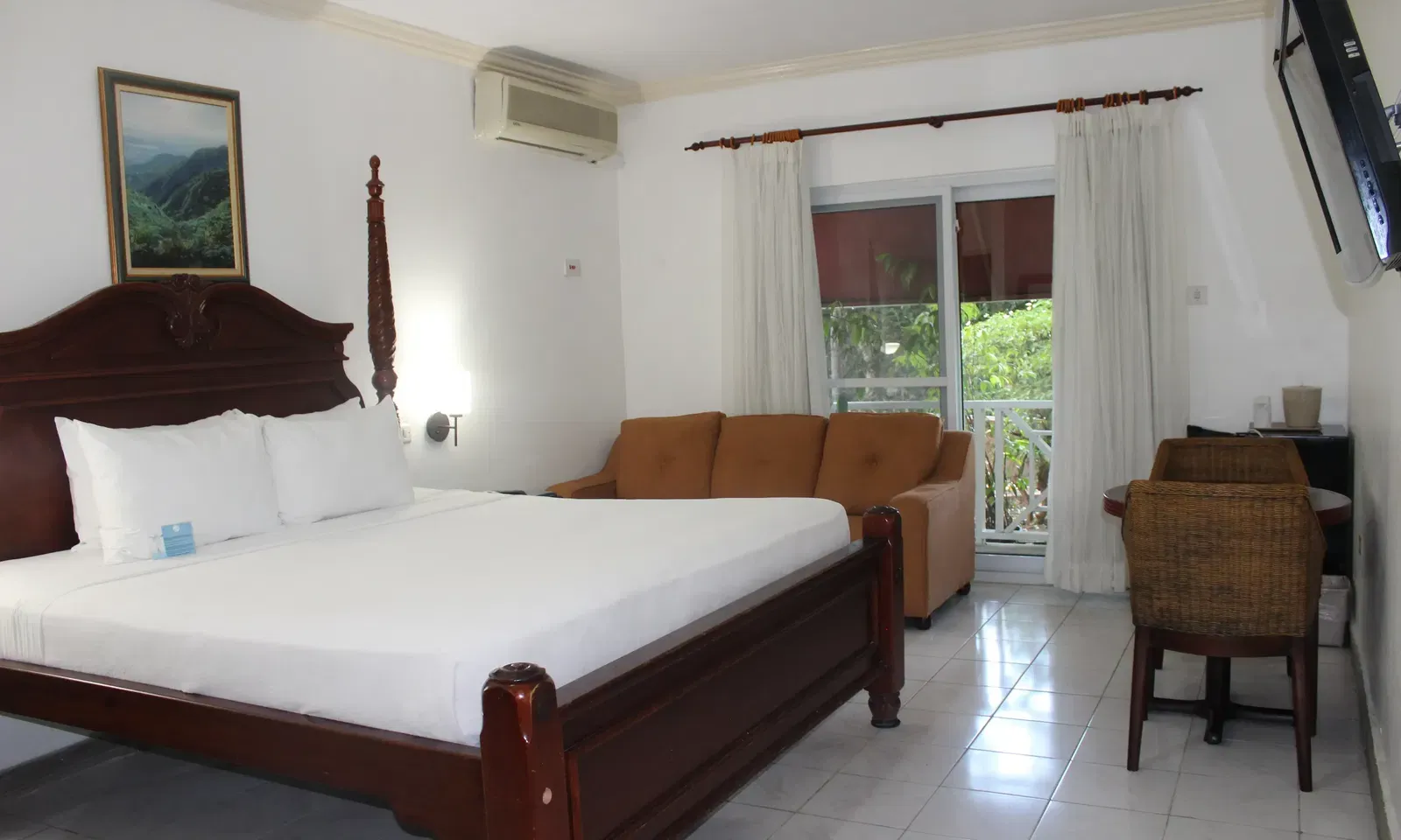 A hotel room features a large wooden bed with white linens, a dark dresser with a television, and a seating area with a couch | Deluxe King Room | Altamont Court, Jamaica