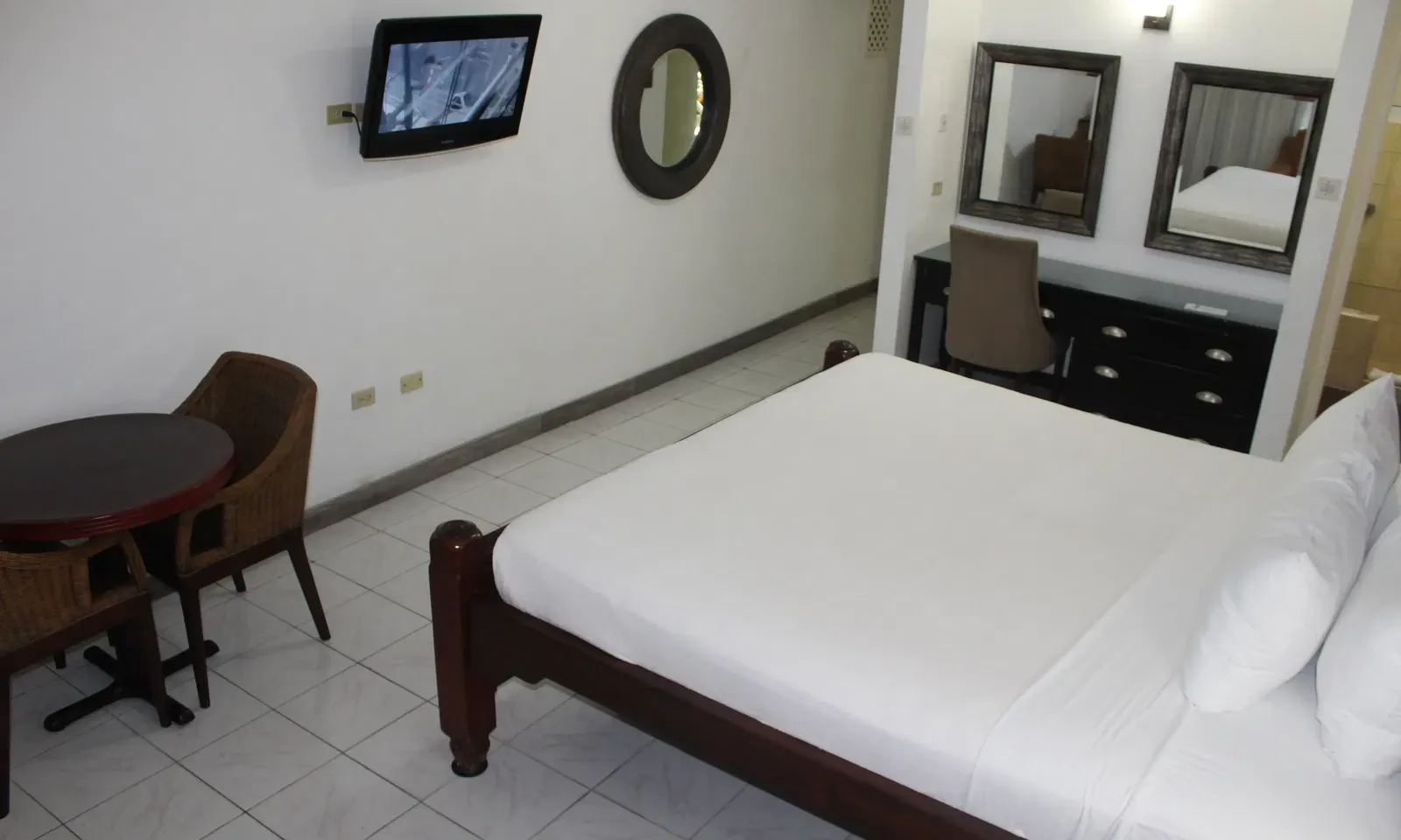 A hotel room presents a bed with white linens, a dark dresser with framed pictures, and a seating area with a table and chairs | Deluxe King Room | Altamont Court, Jamaica