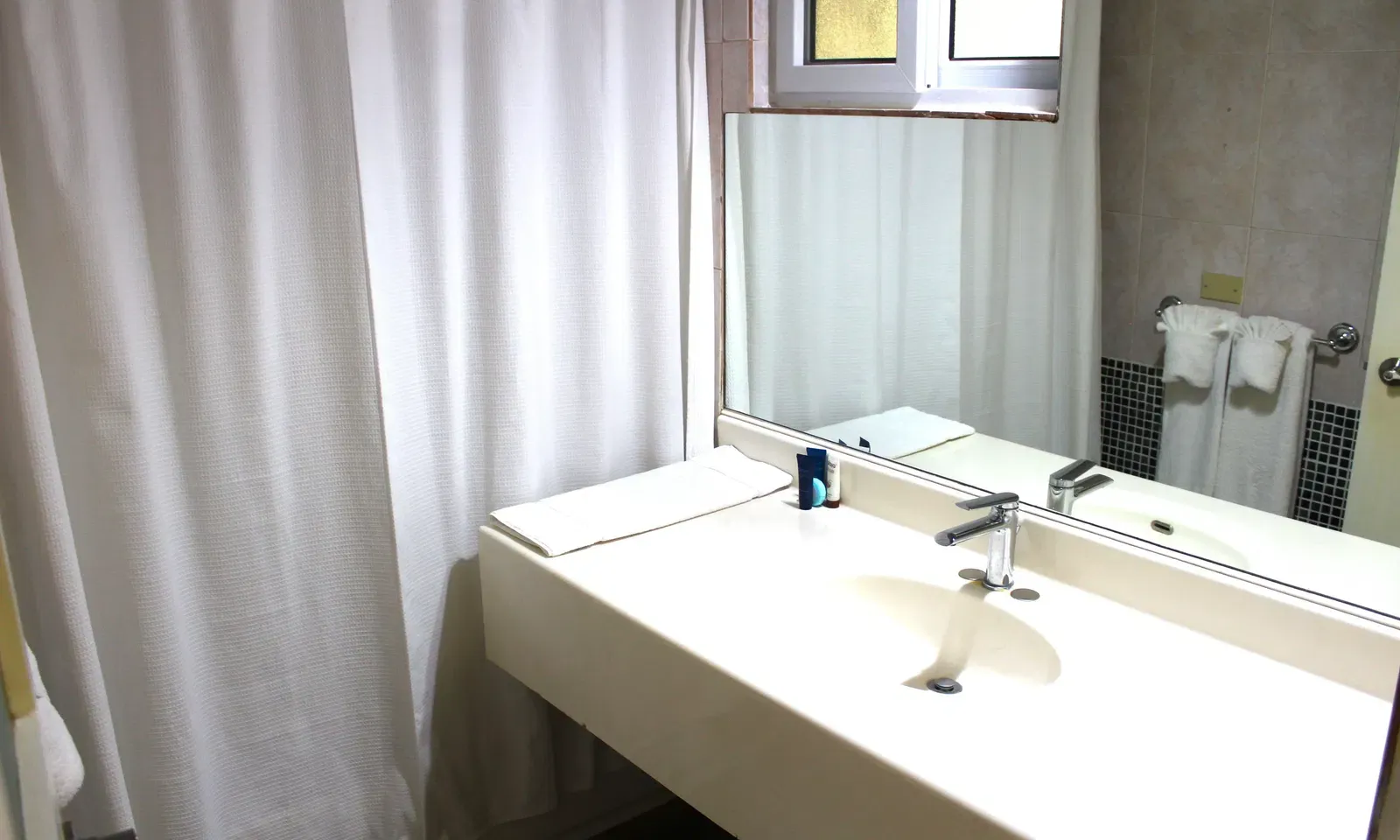 A clean, minimalist bathroom includes a sink with a large mirror and white shower curtains | Executive Double Room | Altamont Court, Jamaica