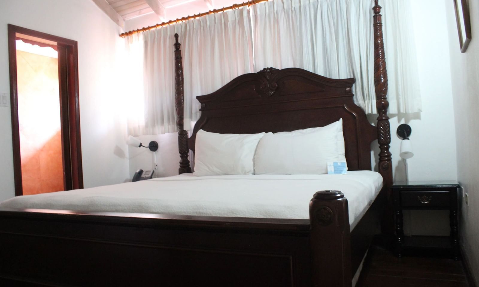 A cozy bedroom is furnished with a dark wooden bed frame, white bedding, and curtains framing a window | Superior Suite | Altamont Court, Jamaica