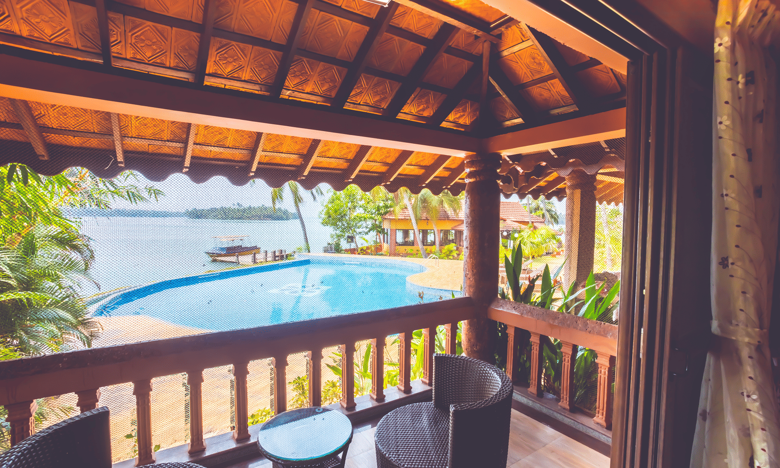 Balcony with tiled roof, two black chairs, and a small table, overlooking water and trees at Paradise Lagoon Resort, Udupi.