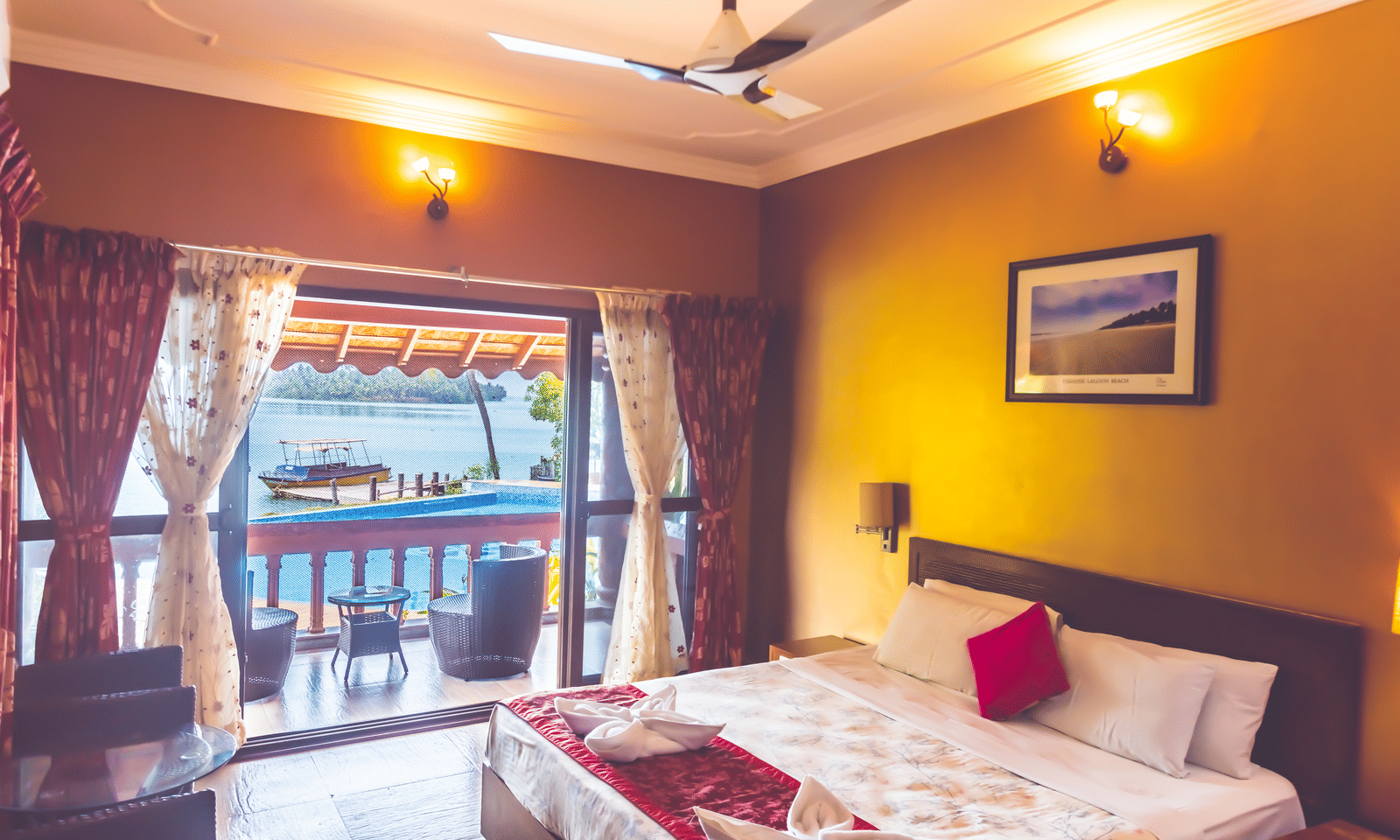 Room with a queen size bed, ceiling fan, framed picture, and view of the river through the window at Paradise Lagoon Resort, Udupi.
