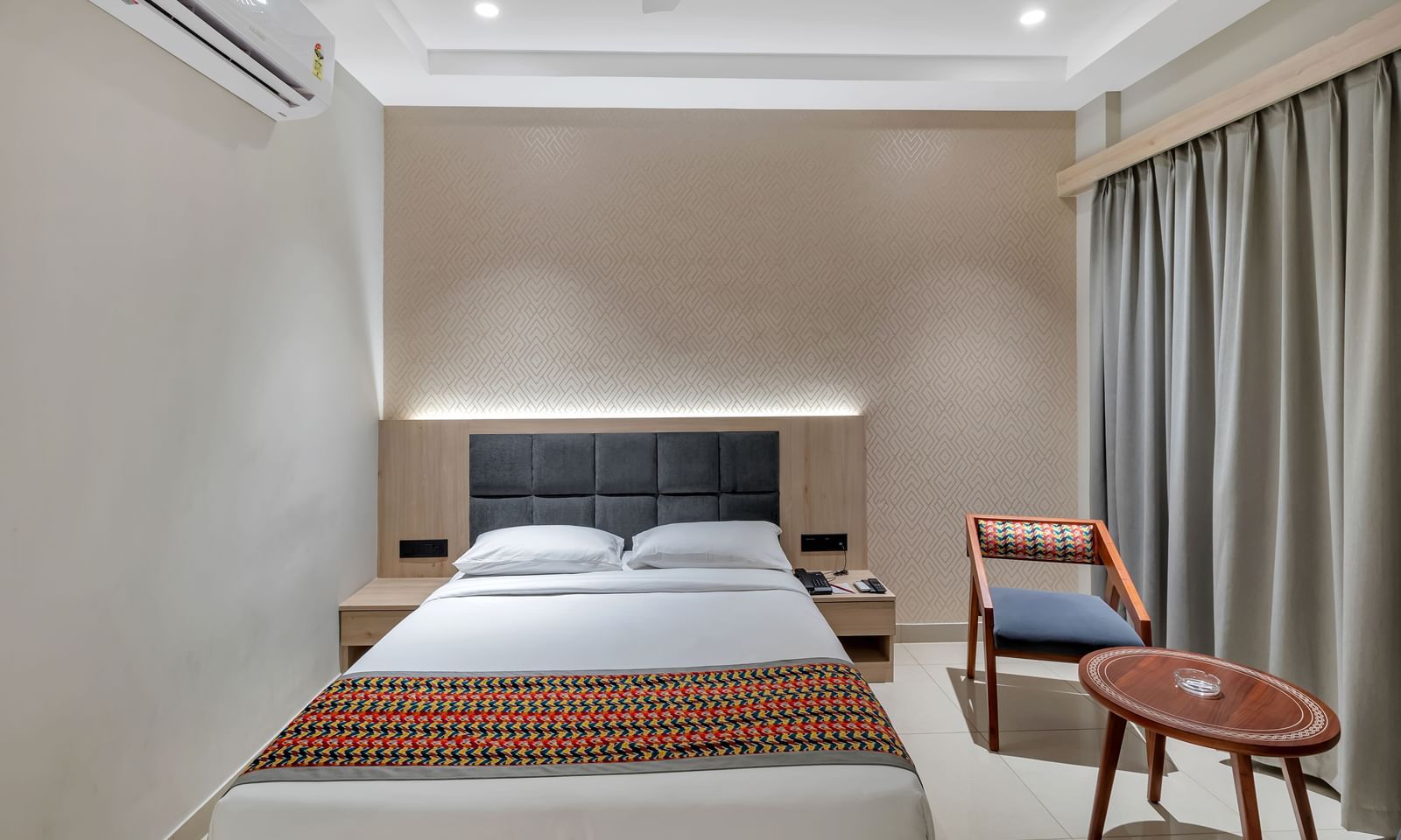 A cosy hotel room with a double bed featuring a colourful patterned throw, bedside tables, a chair, a small round table, and an air conditioning unit | The Citi Residenci Hotel Banskopa