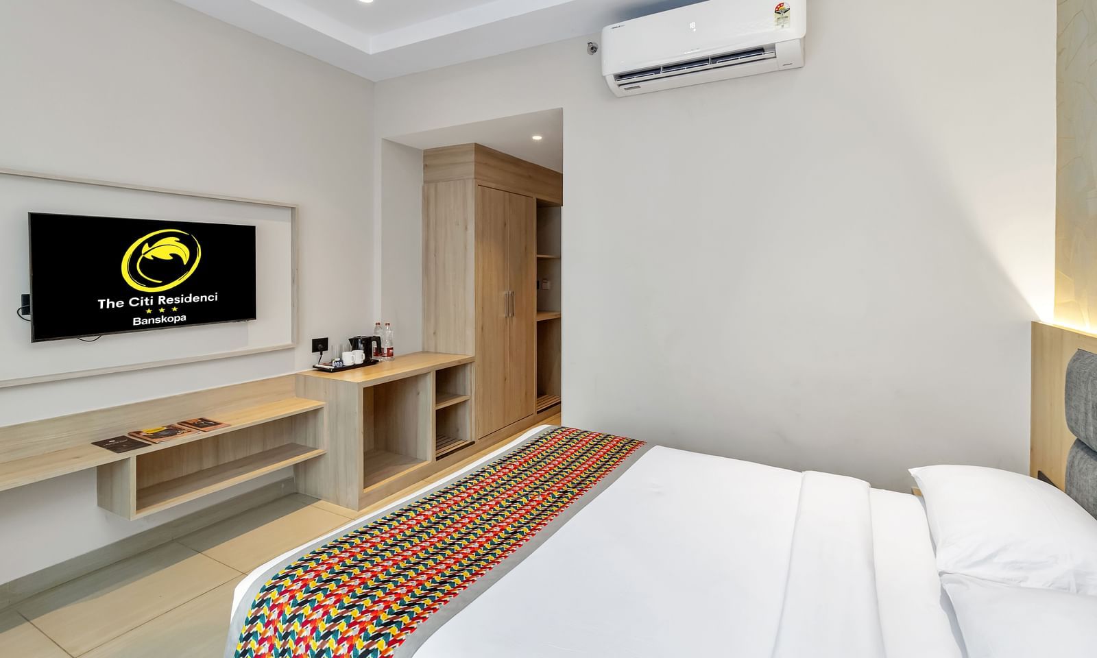 A  room with a bed featuring a colourful patterned runner, a wall-mounted TV displaying a logo, and an air conditioning unit  | The Citi Residenci Hotel Banskopa