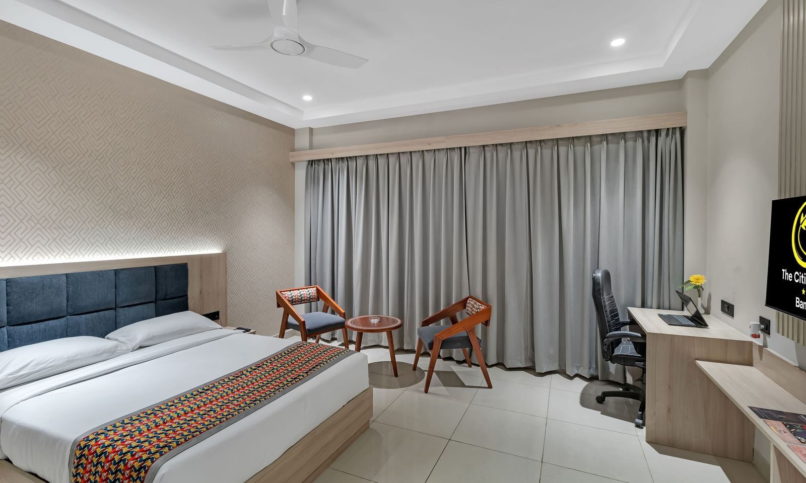 A well-lit hotel room with a king size bed featuring a colourful patterned throw, a seating area with two chairs and a small table, a work desk with a chair, and a television | The Citi Residenci Hotel Banskopa