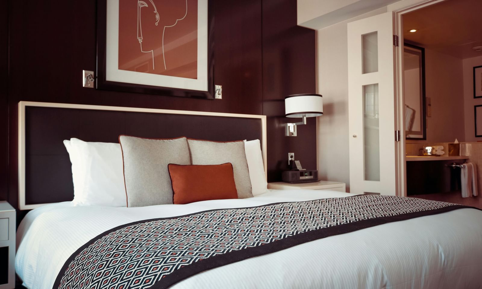 Elegantly decorated hotel bedroom with a large bed, multiple pillows, a patterned bed runner, artwork above the headboard, and a glimpse into an adjoining room.