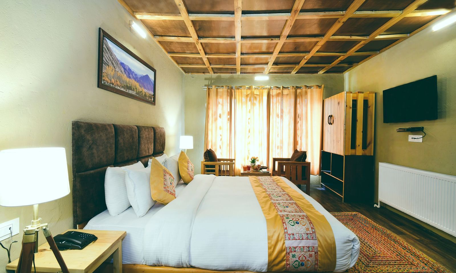 Standard Cottage featuring a king size bed with white linens and a decorative throw, wooden furniture, and a ceiling with a geometric pattern at White Sands Resort, Ladakh.