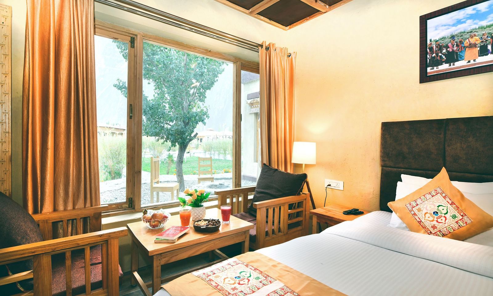 Standard Cottage with a king size bed, a window with orange curtains, a small table and chairs, and a painting on the wall at White Sands Resort, Ladakh.
