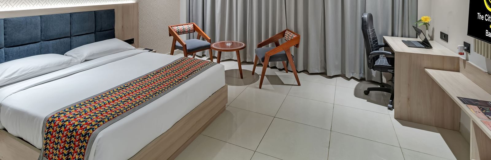 A well-lit hotel room with a king size bed featuring a colourful patterned throw, a seating area with two chairs and a small table, a work desk with a chair, and a television | The Citi Residenci Hotel Banskopa
