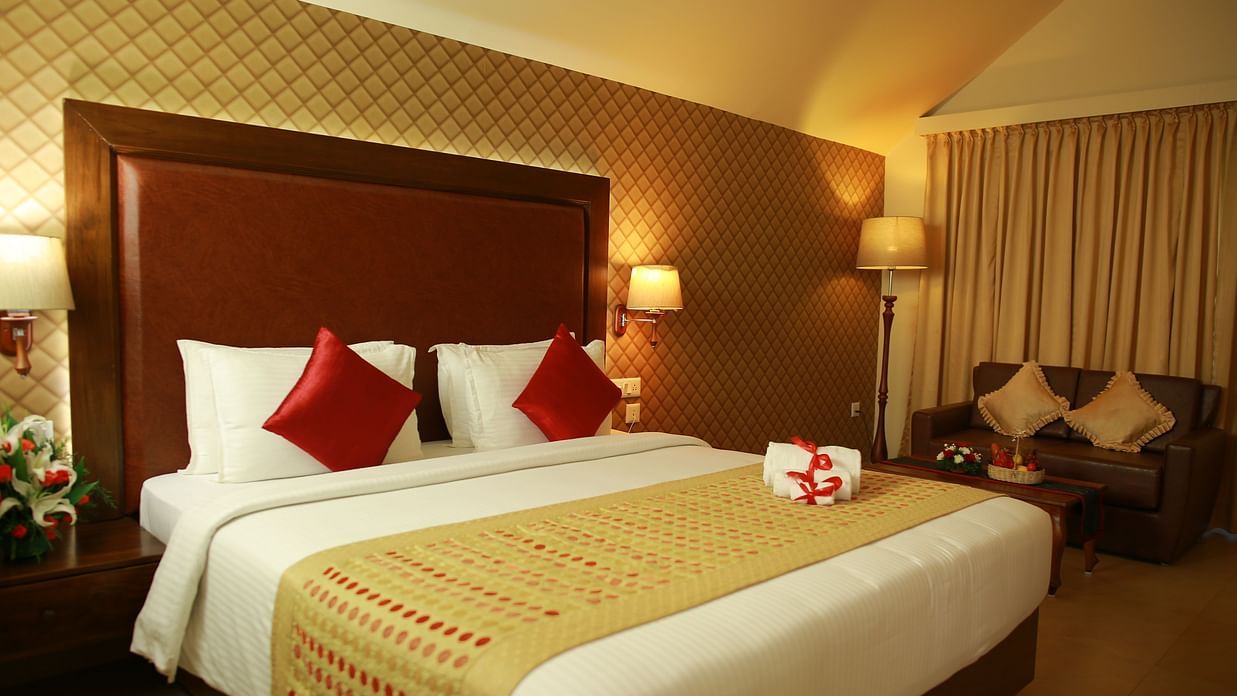 The Atrium Room at Uday Samudra Leisure Beach Hotelfurnished with a large bed featuring white linens, red pillows, and a gold and red patterned runner and includes a seating area with a dark sofa and patterned cushions