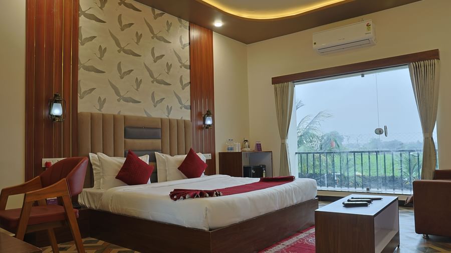 A detailed view of an Executive Room at VITS Select Diamond Resort, Sasan Gir, showing a bed with red pillows, a small sofa, a red chair, and a large window overlooking a field.