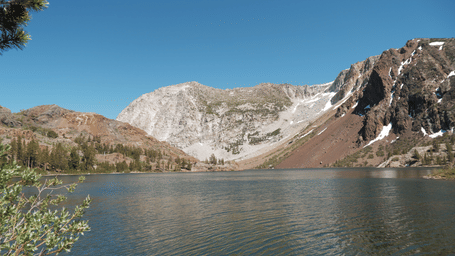A serene panorama of a lake on the foothills of a mountain with surrounding snow-capped peaks under a clear blue sky.