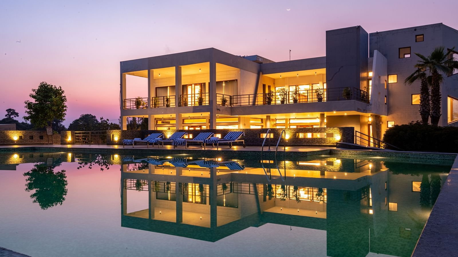 Facade view of DLS Forest Vines Resort, with warm light glowing from its windows and doors, reflected on the shimmering swimming pool beneath a clear sky.