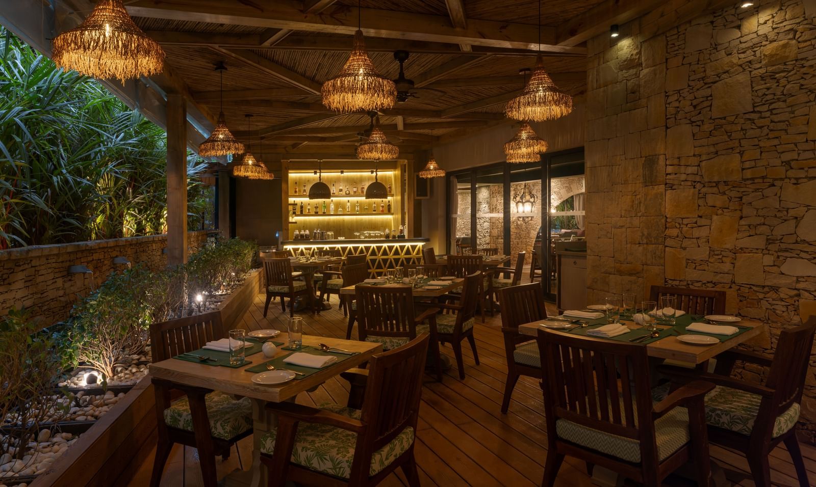 A dimly-lit dining area at the Mila Restaurant with lush green trees on one side at The Evren, Vagator.