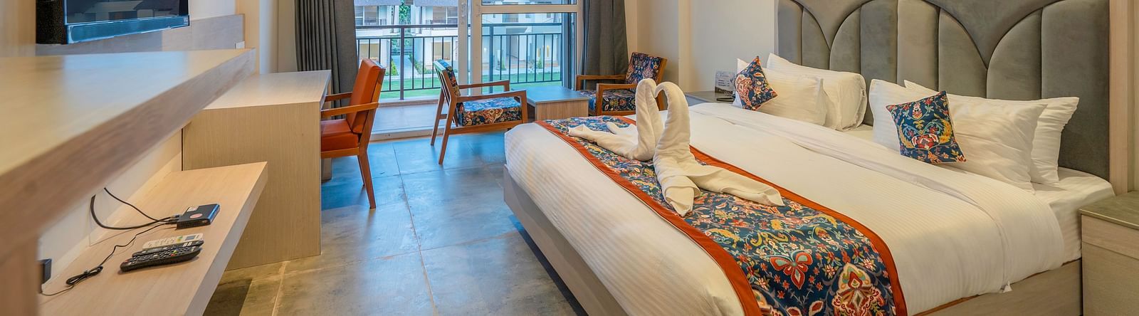A spacious hotel room with a king size bed, a flat-screen TV, a work desk, and a balcony offering a scenic view of the surrounding landscape.