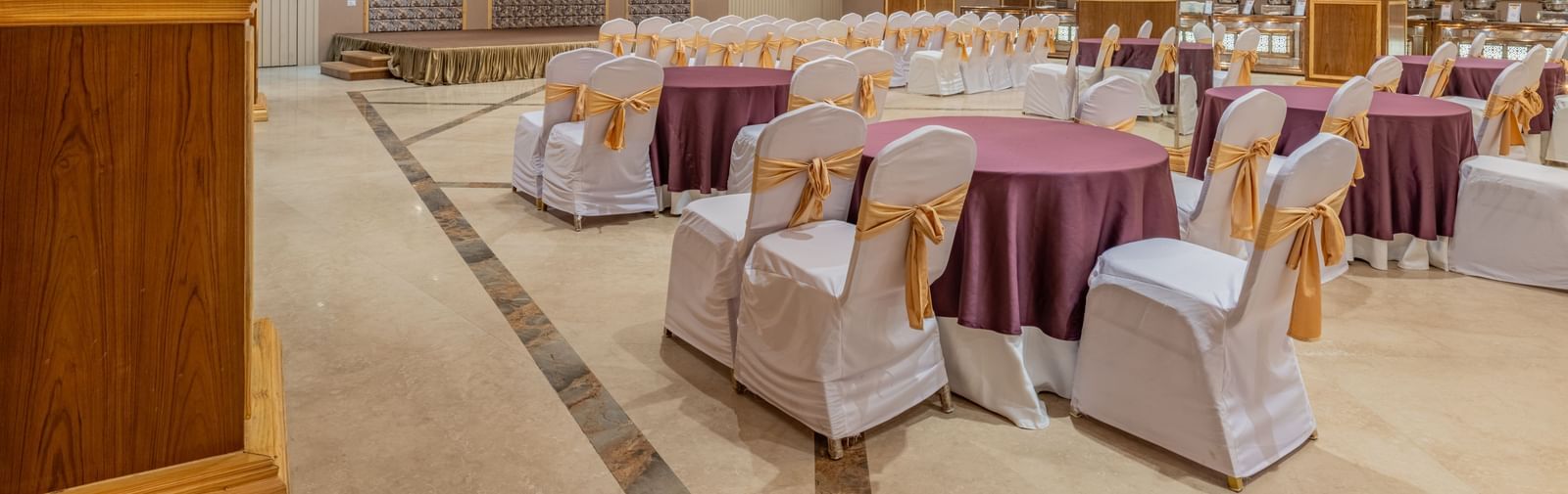 Banquet hall at Hotel Highway King, Bagru, with round tables, decorated chairs, chandeliers, and decorative wall panels.