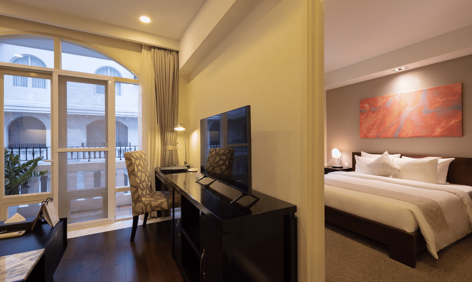 Open plan living area in the Two Bedroom Family Suite at Central Mansions, Phnom Penh with a television, seating area, and adjoining bedroom.