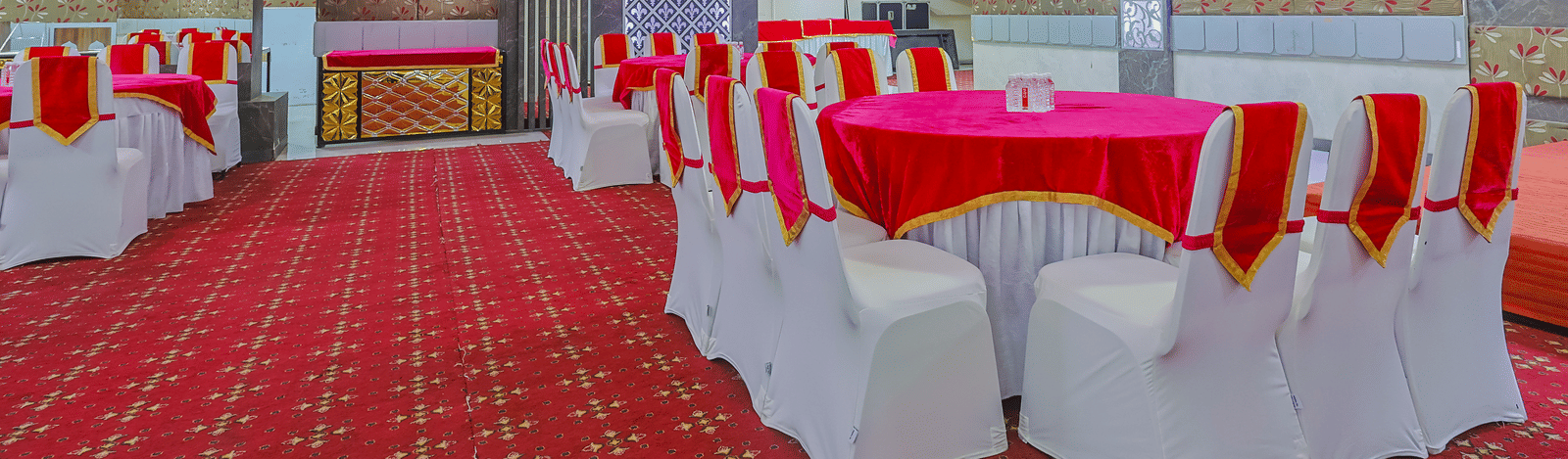  banquet hall set up for an event, featuring tables with white chairs and red tablecloths, arranged on a red patterned carpet.