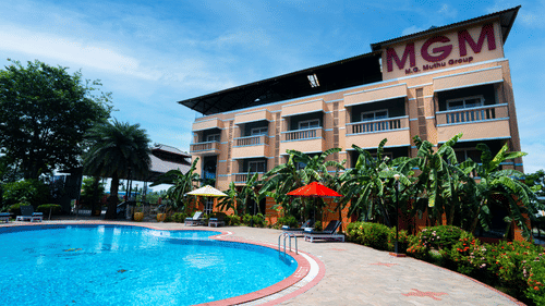 Facade of MGM Vailankanni Residency featuring a large swimming pool on a sunny day surrounded by lawns and bushes