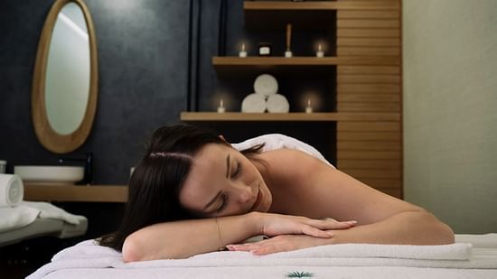 a woman laying on her stomach on a spa bed with the backdrop of towels and essential oils kept in a shelf