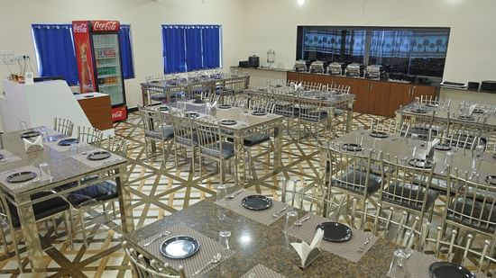 A wide shot of the restaurant at VITS Select Diamond Resort, Sasan Gir. The room is filled with tables and steel chairs, a buffet counter, and a Coca-Cola refrigerator.