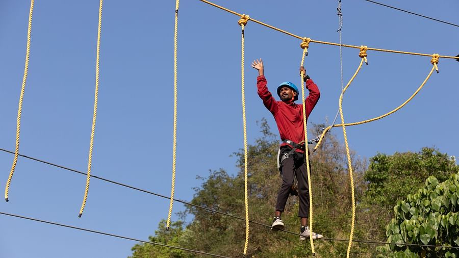 Person navigating a high rope course under a clear blue sky. | Stone Wood Jungle Resort, Dandeli