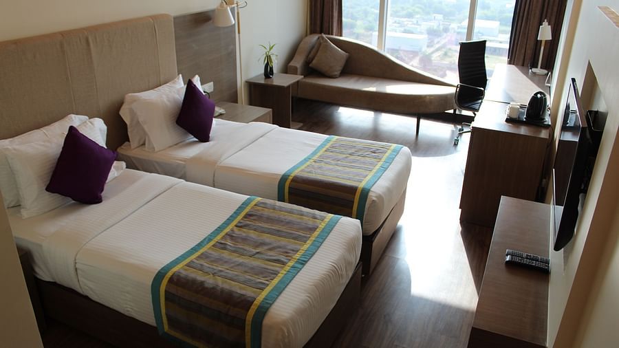 An image showing a pair of twin beds with lush decors, a long sofa with cushion, and a flat screen TV.