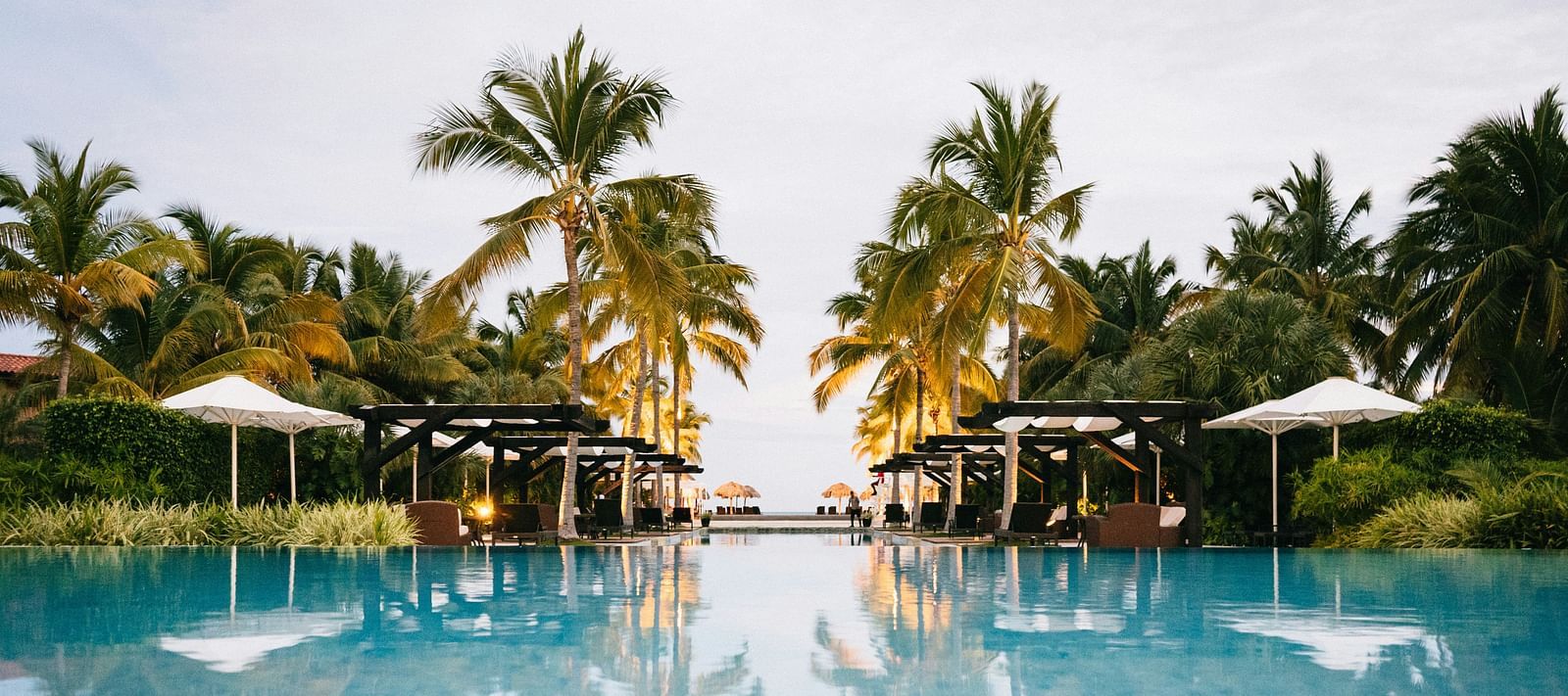 Expansive resort swimming pool reflecting the sky and surrounding palm trees and cabanas.