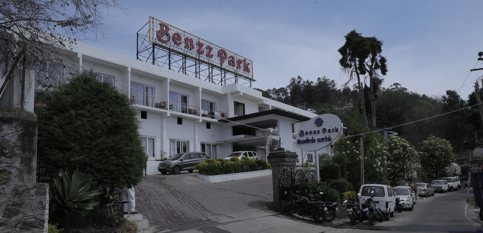 The Benzz Park hotel building exterior on a hillside road, surrounded by trees and vehicles.