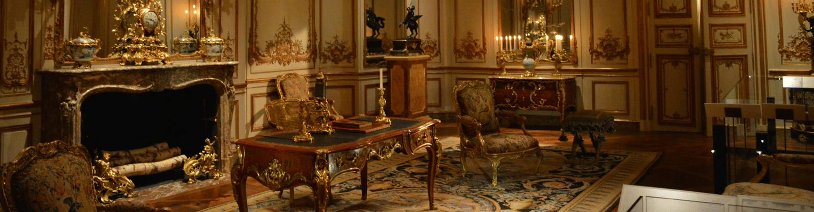 A grand, Baroque style salon with ornate gilded wall panelling, large mirrors, a fireplace, and a crystal chandelier.