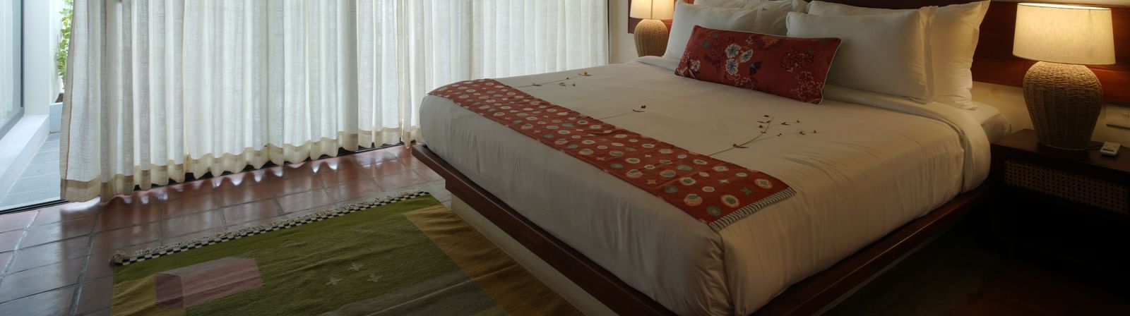 A deluxe room at Vaasta Vagamon featuring a large bed with decorative throw pillows and a patterned runner, and long drapes.