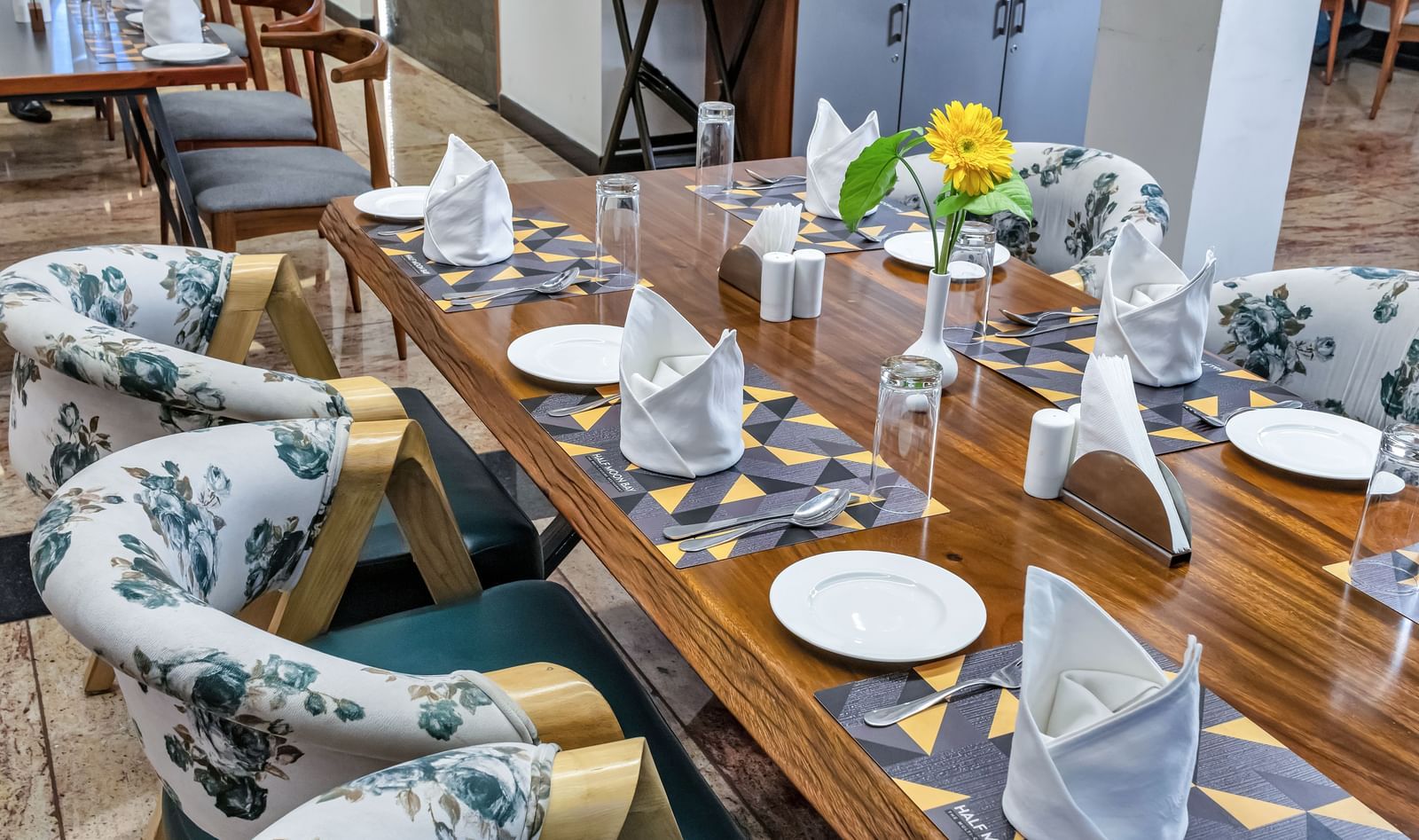 A long wooden dining table set with plates, folded napkins, glasses, and a yellow flower centerpiece, surrounded by chairs with floral patterned upholstery | The Citi Residenci Hotel Banskopa