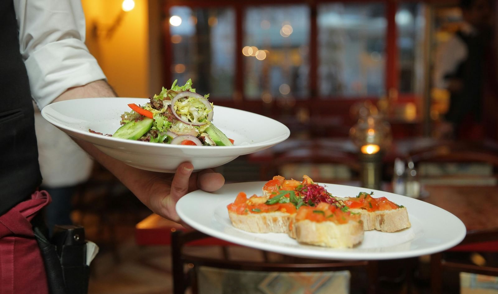 A waiter in a white shirt and black vest carefully carries a plate of salad and a plate of bruschetta in a warmly lit restaurant setting.