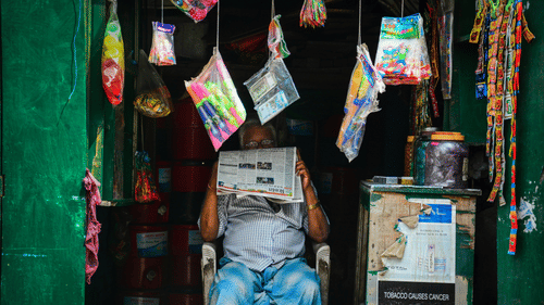 An old man sits in a small shop, reading a paper that covers his face. Bright packs of toys and snacks hang around him. 