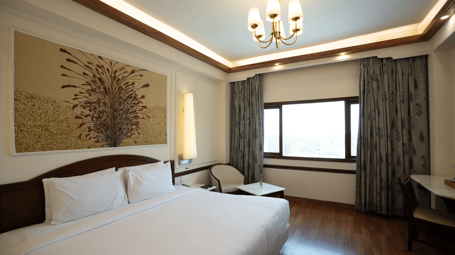 A king size bed with crisp linens sits beneath a floral wall panel and a decorative chandelier in the Deluxe King room at Clarks Amer, Jaipur.