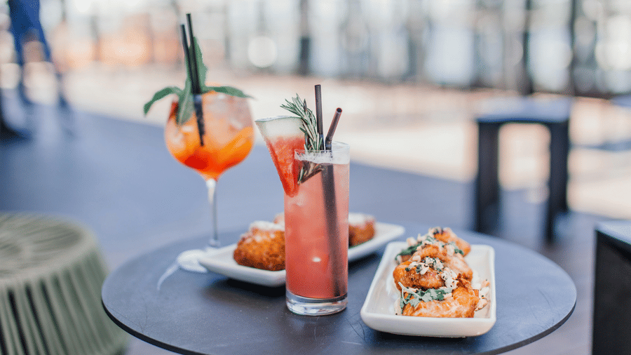 A close up of a small, round, dark outdoor table holding 2 colourful cocktails with garnishes, next to 2 small white plates with snacks