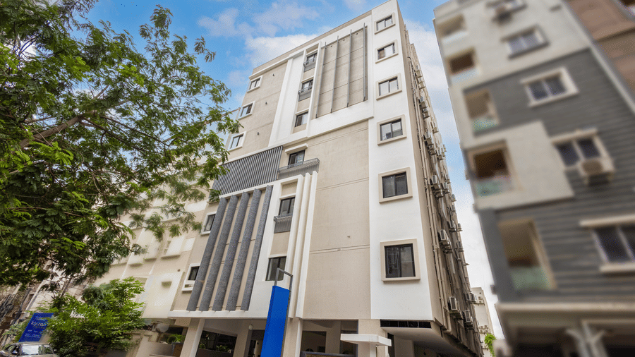 An exterior view showing a multi-storey white building standing between green trees and adjacent urban structures.