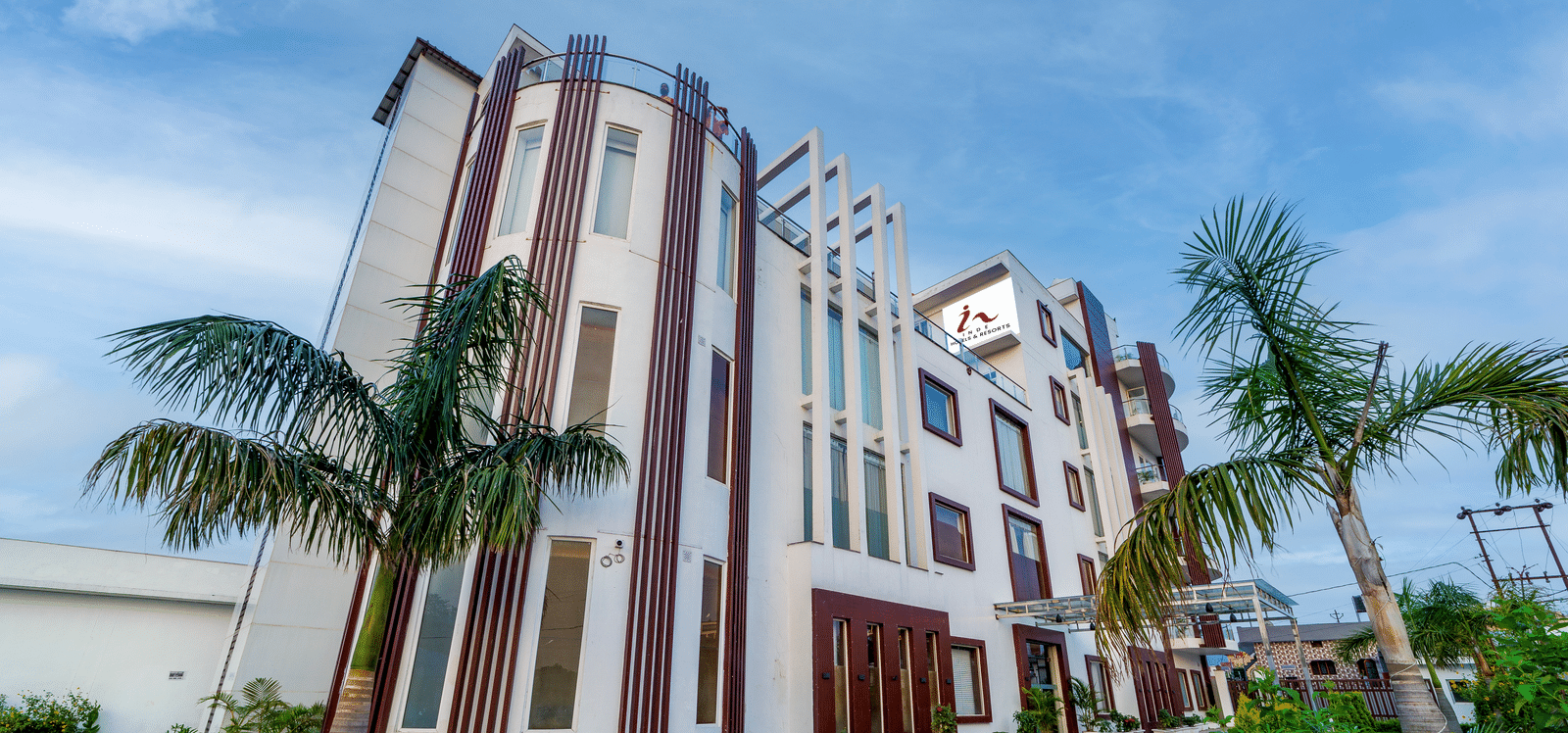 Exterior view of Inde Reva Resorts Rishhikesh showing the multi-storey building with palm trees, paved driveway, and lush green on the right.
