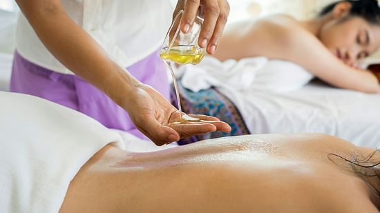 a massage therapist pouring oil on the back of a client as another lies with eyes closed in the background