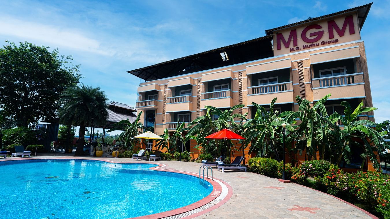 Facade of MGM Vailankanni Residency featuring a large swimming pool on a sunny day surrounded by lawns and bushes