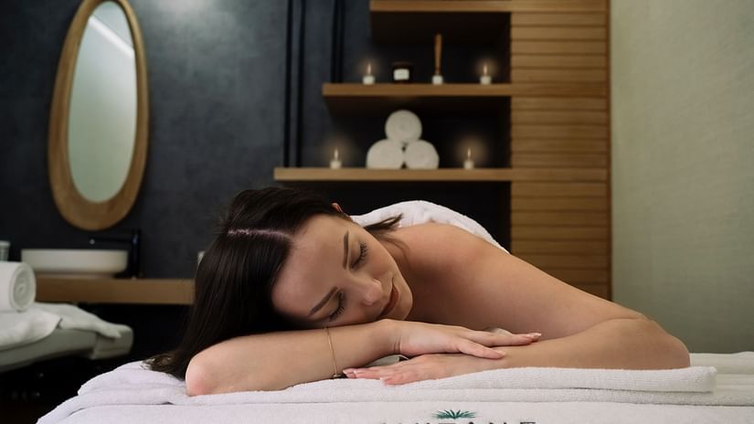 a woman laying on her stomach on a spa bed with the backdrop of towels and essential oils kept in a shelf