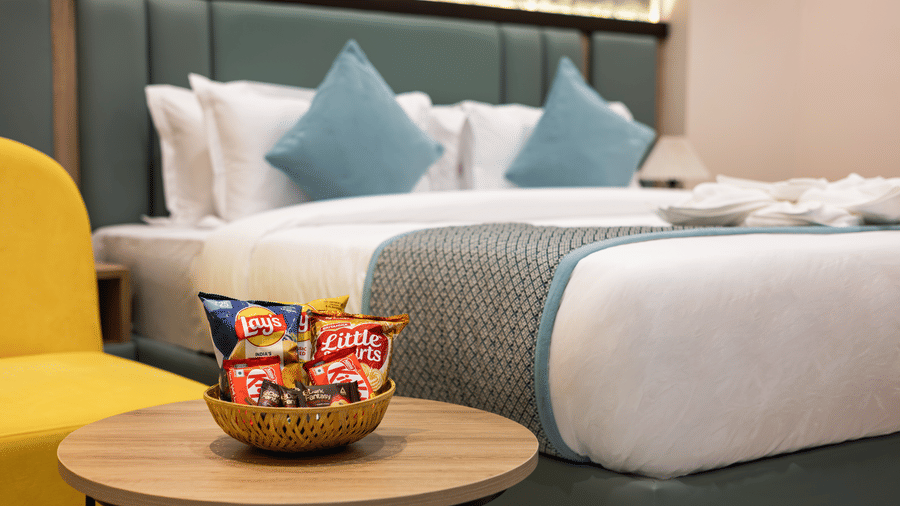 A king size bed with blue accent pillows framed beside a chair, and a small table holding snacks at Monday Hotels & Suites Madhapur Hyderabad.
