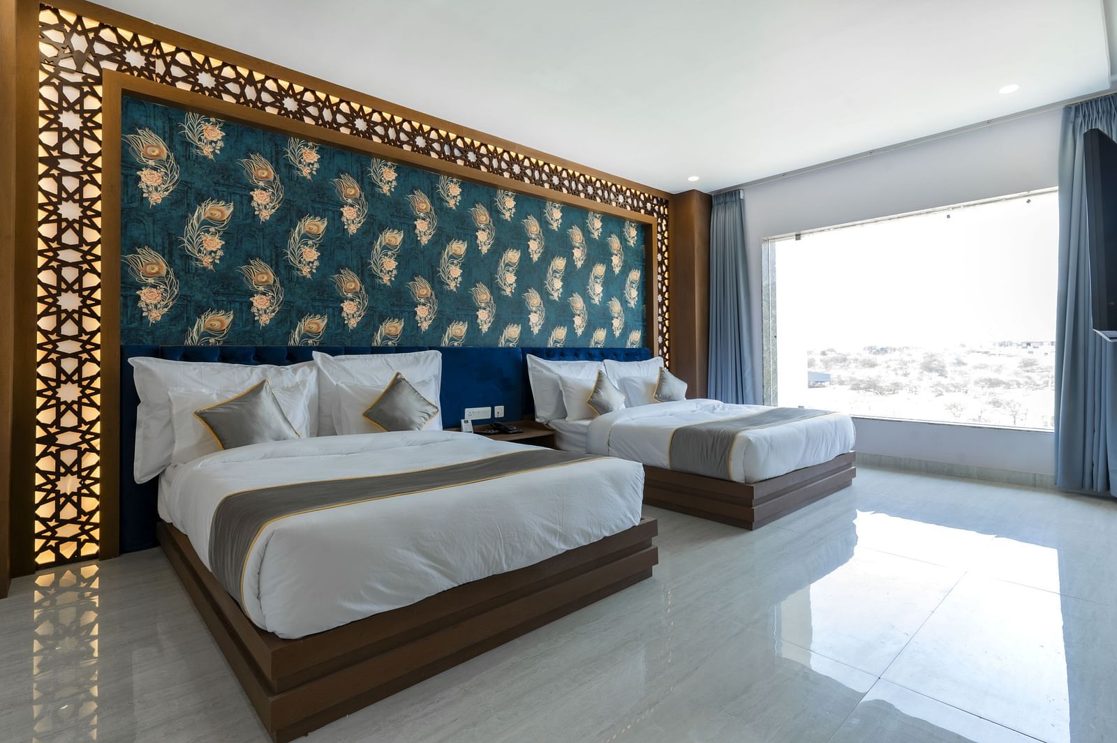 A bedroom at the Bamboo Saa Mulberry Resort featuring two twin beds with white linens and grey throws. A decorative wall panel with a blue and gold pattern is above the beds and a large windows is visible.